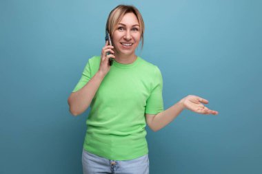 Mavi arka planda modern telefonlu neşeli, tapılası sarışın kadının yatay fotoğrafı..