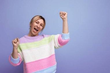 Gülümseyen neşeli, enerjik, sarışın kadın. Mor arka planda sıradan çizgili süveter giyiyor..