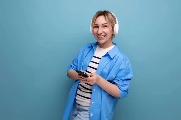 close-up photo of a bright smiling blond young woman dressed in a striped sweater and casual shirt with white wireless headphones communicates in the application in a smartphone on a blue background.