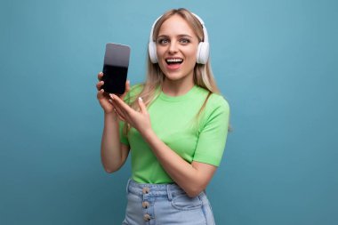 Büyük beyaz kulaklıklı neşeli, enerjik sarışın bir kızın yakın plan fotoğrafı mavi arka planda akıllı telefonun bir taklidini gösteriyor..