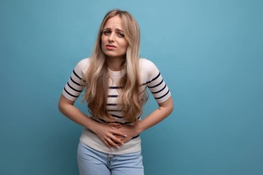 Sıradan giyinmiş mutsuz bir kız, mavi izole bir arka planda hazımsızlık çektiği için karnını sıkıyor..