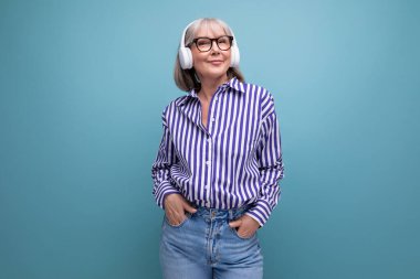 Gri saçlı, gülümseyen, yaşlı bir kadın. Parlak bir stüdyo arka planında kulaklıkla müzik dinliyor..