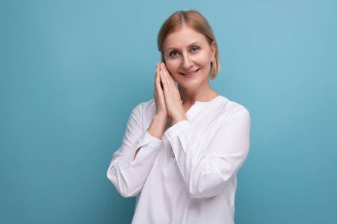 Beyaz gömlekli yetişkin bir kadın stüdyo arka planında gülümsüyor..