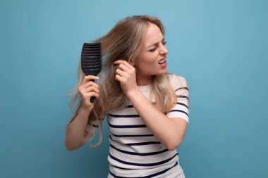 Üzgün, üzgün, sarışın, genç bir kadın. Saçlarını taramakta zorlanıyor. Mavi arka planda düşüyor..