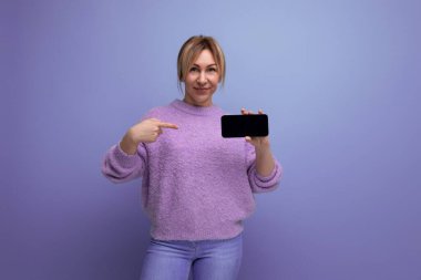 Eflatun kazaklı neşeli sarışın kadın. Mor arka planda akıllı telefon mokaskı ve fotokopi alanı..