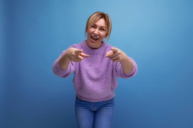 Mor kazaklı, enerjik, sevimli sarışın kadın elinde kamerayla mavi arka planda fotokopi alanı olan bir kadını işaret ediyor..