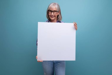 Başarılı gri saçlı, olgun bir kadın parlak stüdyo arka planında mokasen olan boş bir reklam panosu gösteriyor..