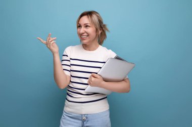 Kendine güvenen, çizgili kazaklı, mavi arka planda dizüstü bilgisayar tutan sarışın bir kadının yakın çekimi..