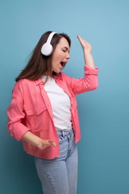 Sırıtan esmer kadın, omuzlarının altında siyah saçlar, gömlek ve kot pantolon içinde kablosuz kulaklıkla müzik dinliyor..