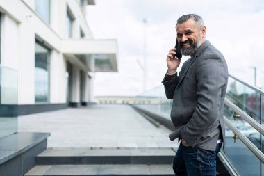 Yetişkin bir erkek patronun yatay fotoğrafı. Gri saçlı ve sakallı, binanın arka planında telefonla konuşan..