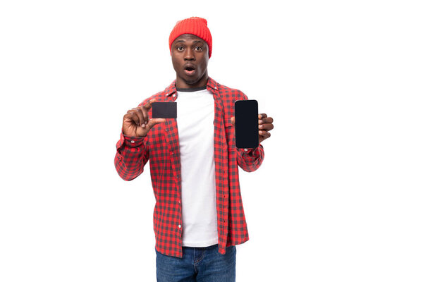 confident young american man in red headdress and shirt showing with hand mockup card and smartphone for shopping on white background with copy space.