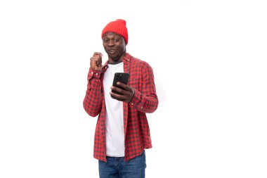 İnternette oturan kırmızı başlıklı yakışıklı Amerikalı genç adam izole edilmiş beyaz arka planda, fotokopi alanı olan akıllı telefon kullanıyor..