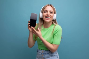 Neşeli ve enerjik sarışın bir kızın resmi. Büyük beyaz kulaklıklı sıradan bir kız. Mavi arka planda akıllı telefonun bir kopyasını gösteriyor..