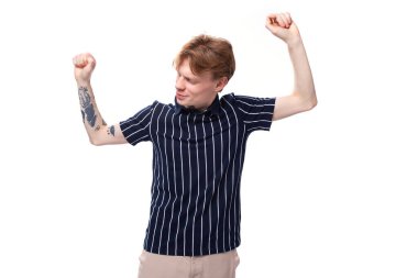 25 'lerinde tişört giymiş, beyaz arka planda dans eden dövmeli sarışın bir adam..