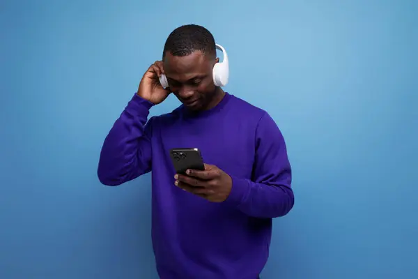 young african man in blue sweater with wireless bluetooth headphones watching video on phone.