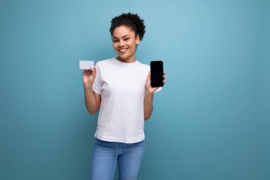 Beyaz tişört giymiş, kıvırcık saçlı, güzel, genç, esmer bir kadın ekranı öne bakan ve plastik kartı olan bir akıllı telefonu dikey olarak tutuyor..