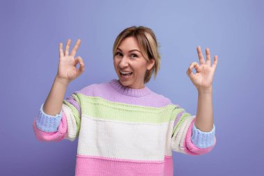 Pastel renkli süveter giyen çekici sarışın kadın mavi arka planda gülümseme sergiliyor..