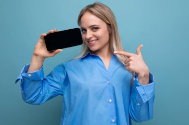 Çekici, kendine güvenen sarışın kız kamerada ekranı olan akıllı bir telefon tutuyor. Mavi arka planda bir web sitesi için boş bir maketi var..