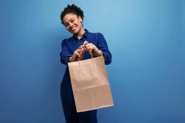young successful woman with dark skin and black curly hair dressed in a blue denim suit holding a craft brown bag.