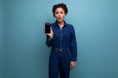 Koyu tenli ve siyah saçlı mavi kot takım elbiseli genç bir kadın akıllı telefon ekranı ve bir maketi gösteriyor..