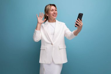 Genç, bakımlı, ince sarışın, beyaz ceketli ve elbiseli kadın akıllı telefon kullanıyor..