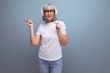 Gri saçlı, modaya uygun, enerjik 60 'lı yaşlarda bir kadın stüdyo arka planında kablosuz kulaklıkla müzik dinliyor..