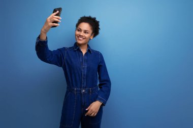 Koyu tenli, siyah kıvırcık saçlı, mavi kot giysili genç bir kadın telefonda selfie çekiyor..