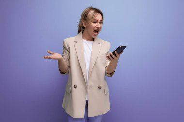 İnançsız ceketli öfkeli sarışın genç bir kadın stüdyo arka planında bir akıllı telefon tutuyor..