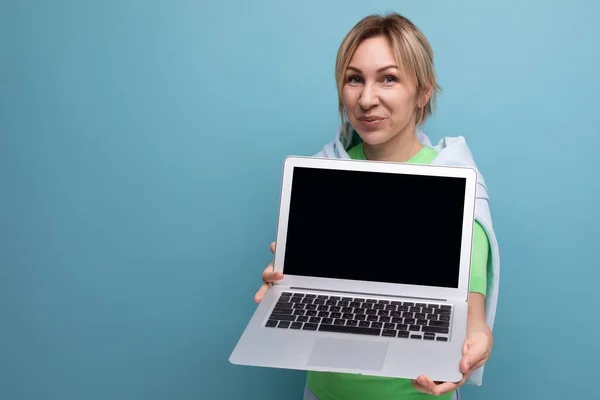 blond positive cute female freelancer in casual outfit showing screen with mockup on laptop on blue background.