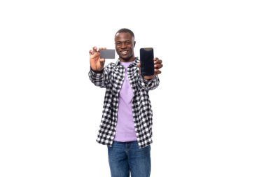 Ekose gömlekli ve kotlu genç esmer adam kredi kartı ve akıllı telefonu fotokopi alanı ile izole edilmiş beyaz arka planda gösteriyor..