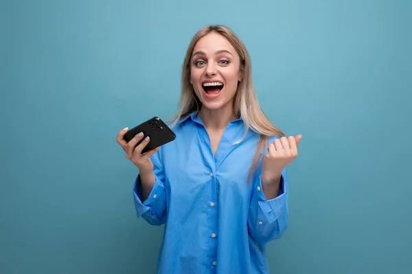 lucky charming blonde girl smiling holding smartphone in hands on isolated blue background.