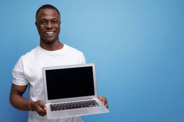 Beyaz tişörtlü başarılı genç Afrikalı adam mavi arka planda dizüstü bilgisayar tutuyor..