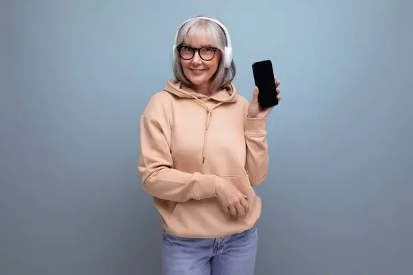 60s mature woman with gray hair with a smartphone in her hands is studying digital technologies in headphones on a studio background with copy space.