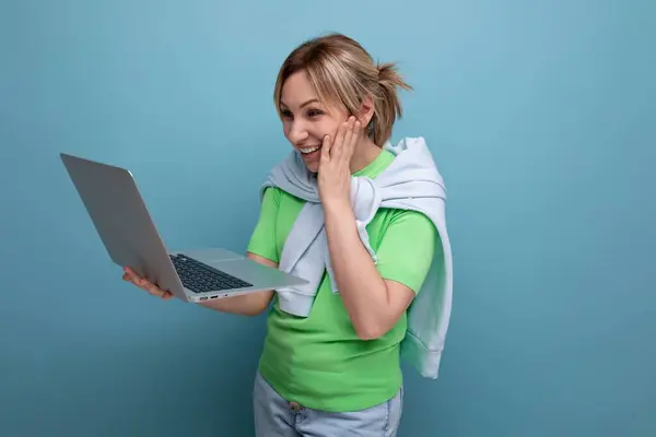 young pensive woman in casual attire working using a laptop in her hands on a blue background.
