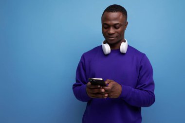 Mavi eşofmanlı, kablosuz kulaklıklı ve mavi arka planlı akıllı telefonlu yakışıklı bir Amerikalı..