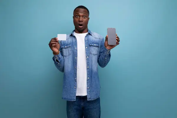 handsome surprised young attractive african man dressed casually in denim clothes holding credit card and smart phone on background with copy space.