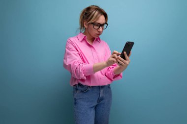 Pembe gömlekli, kot pantolonlu ve gözlüklü, at kuyruklu, sarışın, başarılı bir ofis çalışanı. Akıllı telefon kullanıyor..