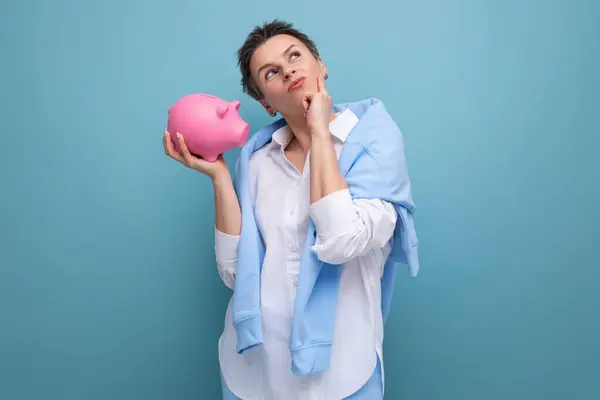 a pensive young woman with a short haircut holds a piggy bank and thinks where to invest the accumulated money.