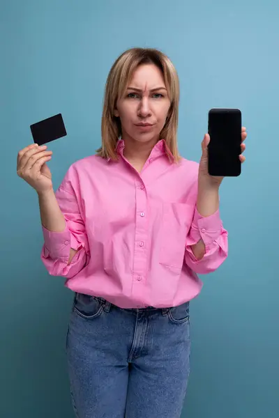 young well-groomed blond caucasian woman with flowing hair is dressed in a pink blouse and jeans holding a smartphone and a credit card.