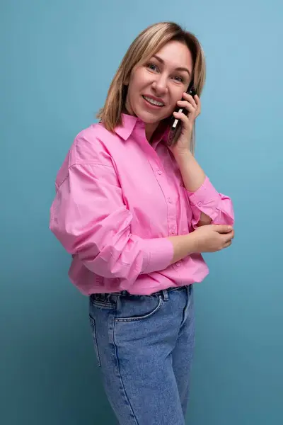 young well-groomed blond caucasian woman with flowing hair is dressed in a pink blouse and jeans talking on the phone.