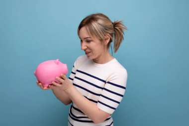 Çekici bir genç kadının yatay fotoğrafı. Günlük kıyafetler içinde. Mavi arka planda bir kumbara dolusu para ve fotokopi alanı..