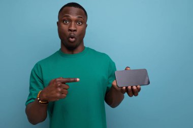Kısa saçlı, basit bir tişört giymiş genç, şaşırmış Afrikalı bir adam elinde arka planda fotokopi alanı olan bir akıllı telefon tutuyor..