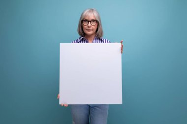 Başarılı gri saçlı, olgun bir kadın parlak stüdyo arka planında mokasen olan boş bir reklam panosu gösteriyor..