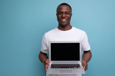 Beyaz tişörtlü başarılı genç Afrikalı adam mavi arka planda dizüstü bilgisayar tutuyor..