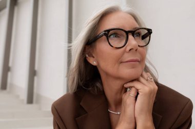 Gözlüklü, gri saçlı, kendinden emin yaşlı bir kadının portresi binanın arka planında kahverengi bir ceket giyiyor..