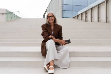 Gri saçlı, kahverengi ceketli, kendine güvenen yaşlı bir kadın bir iş binasının merdivenlerinde oturuyor..