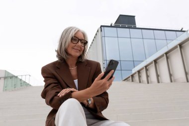 Kısa saçlı, kısa saçlı, ceketli ve pantolonlu orta yaşlı bir kadın şehirde bir iş merkezinin ve ofislerin arka planına doğru yürüyor..