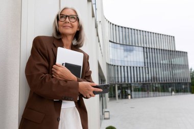 Güzel, başarılı, olgun, gri saçlı, elinde evrakları olan bir iş kadını bir iş merkezinin arka planında duruyor..