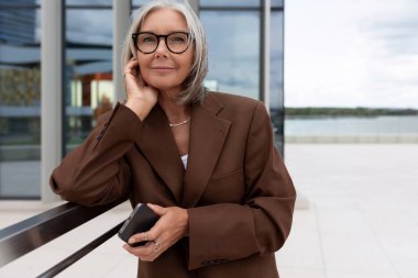 Gri saçlı, kahverengi ceketli, kendine güvenen yaşlı kadın sokakta bir toplantı bekliyor..