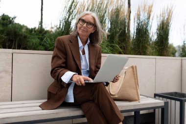 Yetişkin bir iş kadını zarif kahverengi bir ceket, pantolon ve gömlekle şehir caddesinde yürüyor..